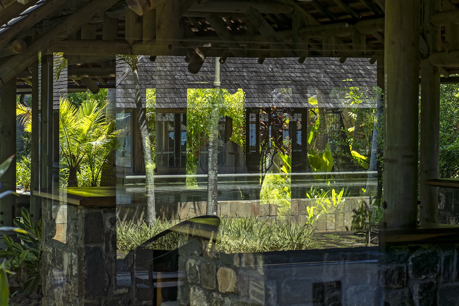 Vue à travers une pergola en bois à colonnes de pierre vers un bâtiment à toit d'ardoise, végétation tropicale