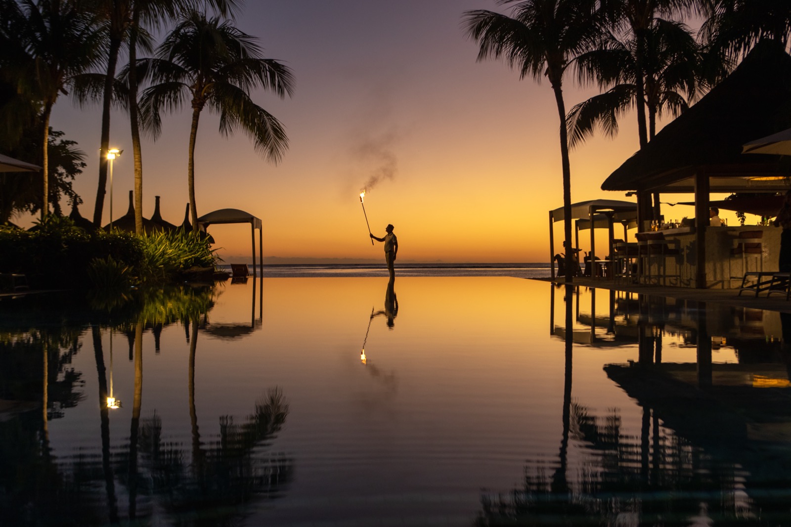 Un employé en silhouette tient un long flambeau allumé au bord d'une piscine à débordement