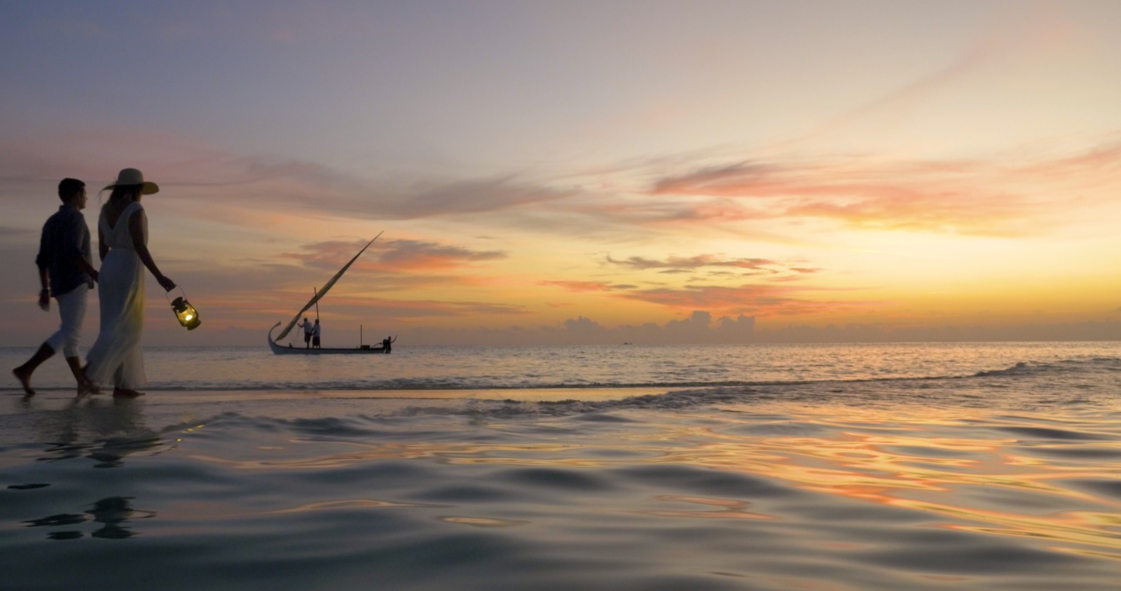 Un couple marche pieds nus au bord de l'eau sous un coucher de soleil flamboyant, la femme au chapeau à larges bords tenant une lanterne allumée, un dhow à l'horizon