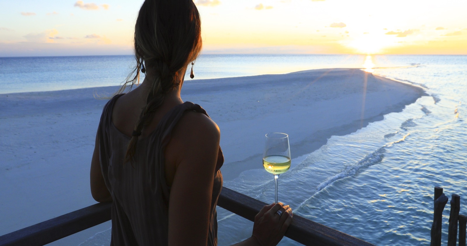 Une femme de dos tient un verre de vin blanc et contemple un banc de sable au coucher du soleil depuis une terrasse en bois sur l'eau