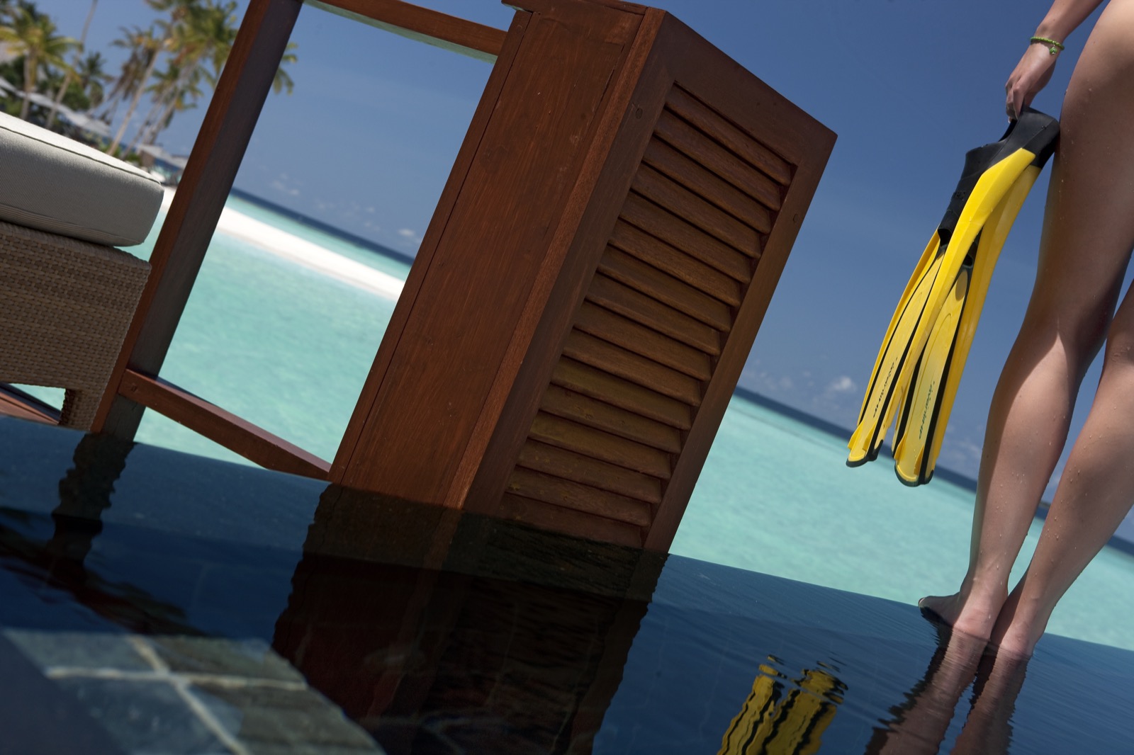 Une femme tient une paire de palmes de plongée jaune vif au bord d'un deck en pierre sombre surplombant un lagon turquoise