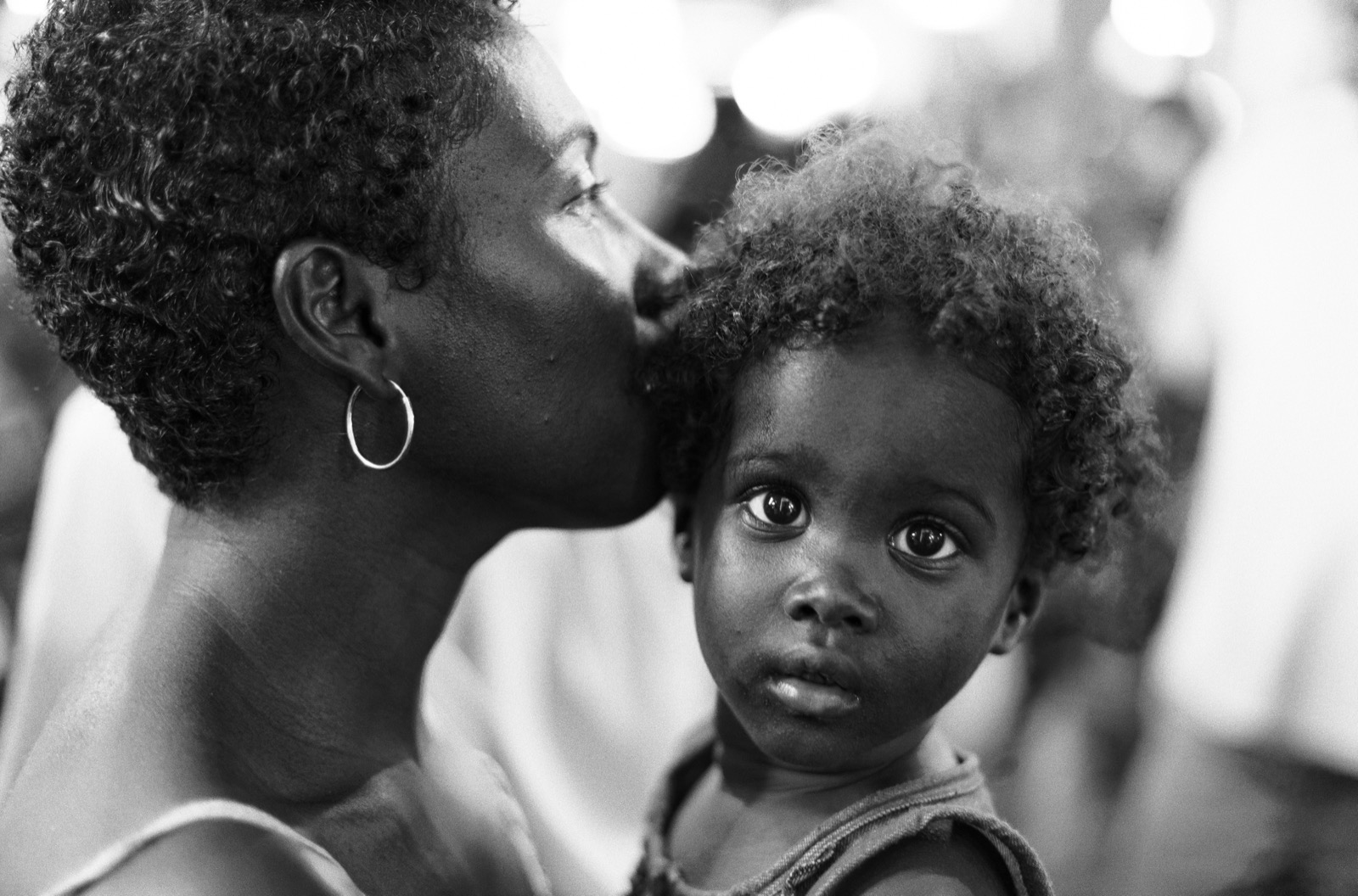 Mère embrassant le front de son enfant qui regarde l'objectif, photographie en noir et blanc