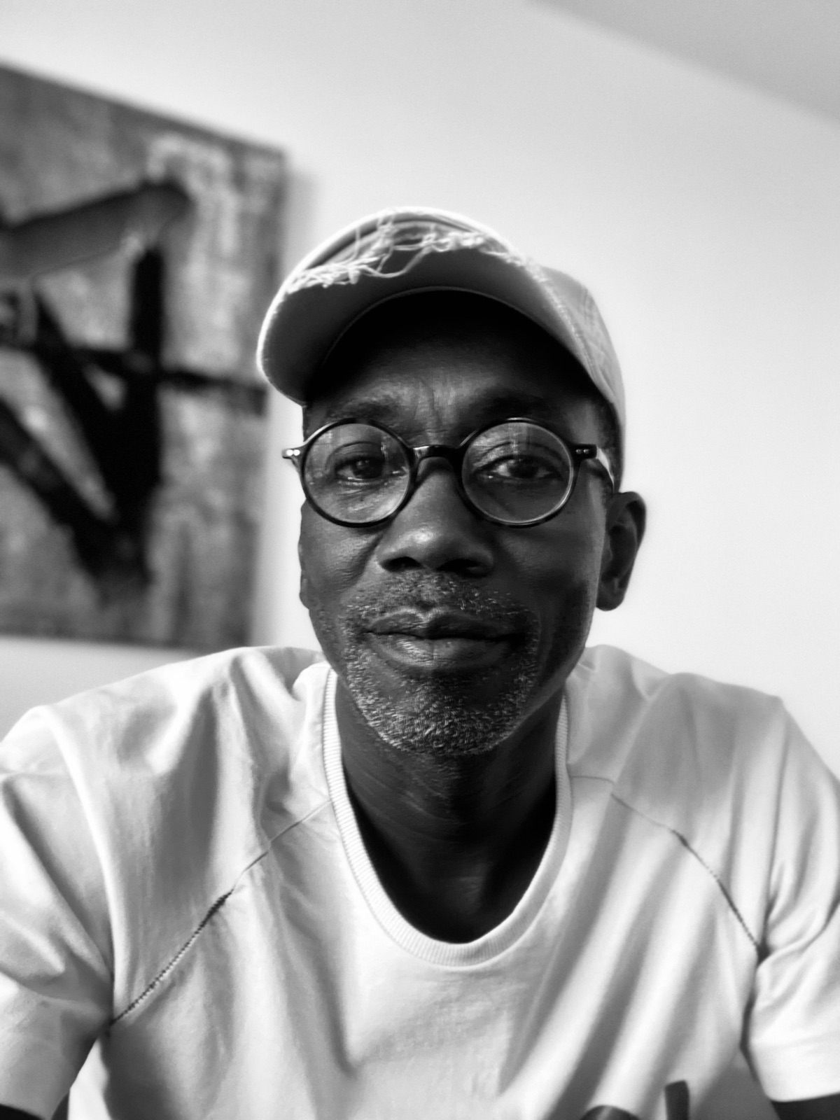 Autoportrait de Bamba Sourang, casquette et lunettes rondes, devant l'une de ses œuvres, photographie en noir et blanc