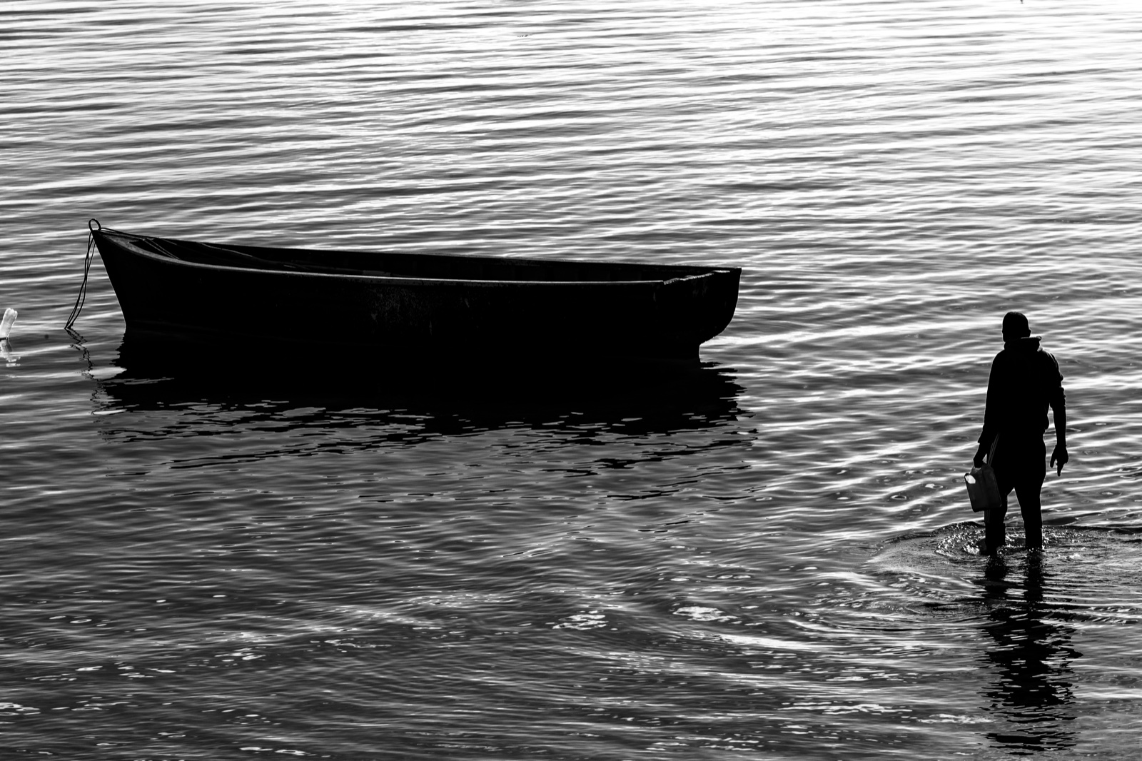 Silhouette d'un homme marchant dans l'eau vers une barque en bois, photographie en noir et blanc